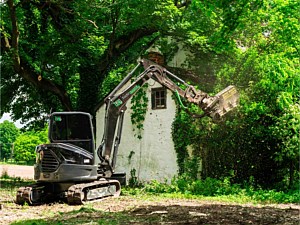 Excavator with forestry mulcher working around abandoned farm house in green lane